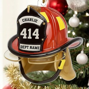 May include: A red and gold firefighter helmet Christmas ornament. The ornament has a black shield with the name "CHARLIE" and the number "414". The words "DEPT NAME" are also on the shield. The ornament is hanging on a Christmas tree.