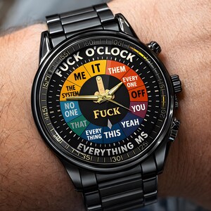 May include: Black wristwatch with a round face featuring the text "FUCK O'CLOCK" and various phrases like "ME," "THEM," and "YOU." The watch has a black band and gold-colored hands.