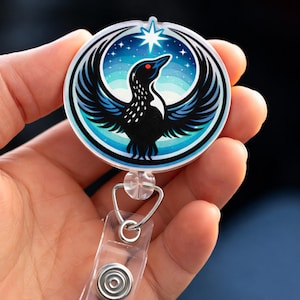 May include: A circular badge reel featuring a black bird design with a red eye, set against a blue and white starry sky. The bird has outstretched wings. The reel has a clear plastic strap and a metal clip.