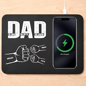 May include: Black wireless charging pad with the word "DAD" in white, along with the phrases "THE MAN, THE MYTH, THE LEGEND." Below are illustrations of fists bumping, with names "WILLIAM", "HERNY", and "LUCIA." A phone is charging at 67%.