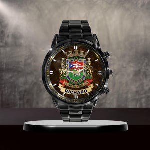 May include: A black wristwatch with a brown face featuring a coat of arms design and the name "RICHARD" on a banner. The watch has a black metal band and is displayed on a white stand.
