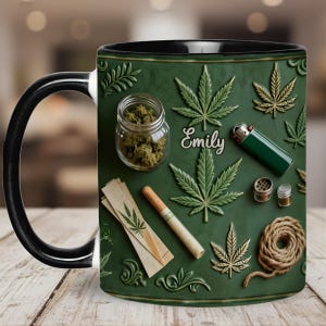 May include: A ceramic mug with a black handle and rim, featuring a green background with cannabis leaf designs and related items. The mug includes a jar of cannabis, a lighter, and the name "Emily".