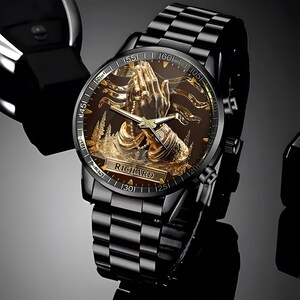 Puede incluir: Reloj de pulsera negro con una esfera dorada detallada que representa manos rezando, una escena de bosque y el nombre "RICHARD". El reloj tiene una correa de metal negro y un segundero dorado.