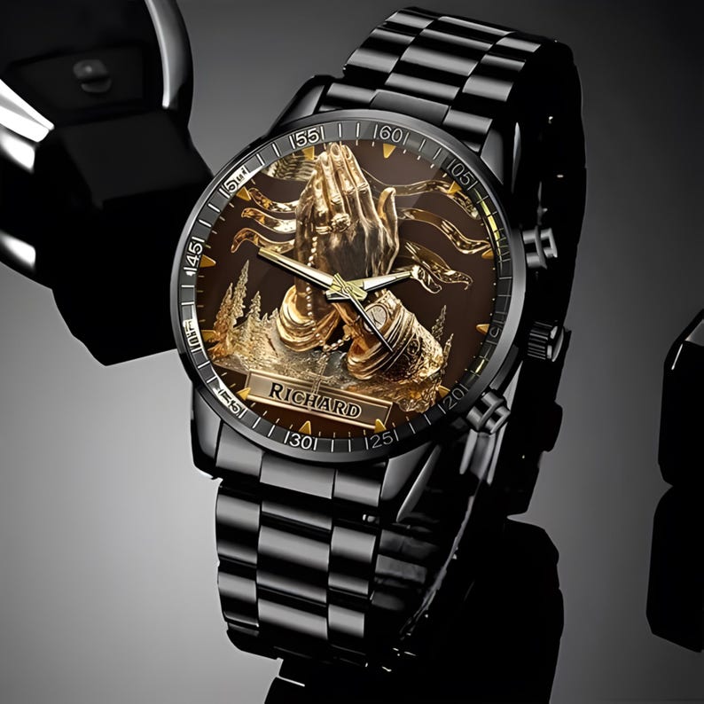 Puede incluir: Reloj de pulsera negro con una esfera dorada detallada que representa manos rezando, una escena de bosque y el nombre "RICHARD". El reloj tiene una correa de metal negro y un segundero dorado.