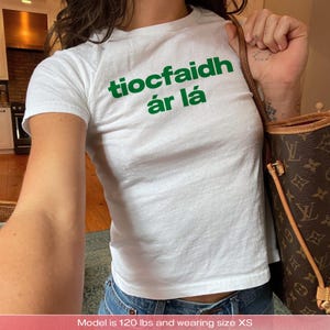 以下が含まれることがあります： 緑色の「tiocfaidh ár lá」の文字が入った白いTシャツ。半袖のトップスは綿のようです。モデルは青いジーンズを履き、茶色のハンドバッグを持っています。テキストはアイルランド語です。