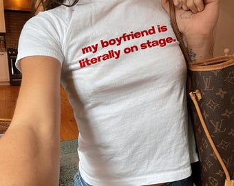 Könnte beinhalten: Weißes T-Shirt mit dem roten Text "my boyfriend is literally on stage." Das Shirt hat einen Rundhalsausschnitt und wird mit einer blauen Jeans kombiniert. Eine braune Handtasche ist im Hintergrund zu sehen.