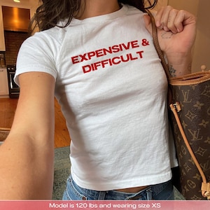Könnte beinhalten: Weißes T-Shirt mit dem roten Text "EXPENSIVE & DIFFICULT". Das Shirt hat kurze Ärmel und einen Rundhalsausschnitt. Das Model trägt eine blaue Jeans und trägt eine braune und beige Handtasche.