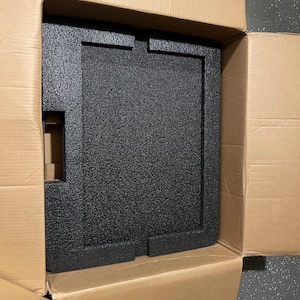 Könnte beinhalten: Ein schwarzer, strukturierter Schaumstoffeinsatz befindet sich in einem Karton. Der Einsatz hat eine rechteckige Form mit Aussparungen, wahrscheinlich zum Schutz eines Artikels beim Versand. Die Box ist geöffnet und zeigt den Inhalt.