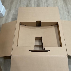 May include: An open, unfolded cardboard box with a rectangular cutout in the center. The box is brown and has flaps extending outward. The cutout has a handle-like feature at the bottom. The box is on a light-colored wooden floor.