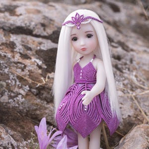 Puede incluir: Una muñeca con pelo largo blanco y un vestido, diadema y sandalias de macramé morado. La muñeca está de pie entre rocas y flores moradas. El vestido tiene un diseño único en forma de pluma y está adornado con pequeñas cuentas blancas.