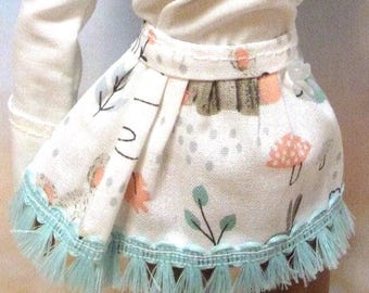 May include: A doll with dark skin and curly black hair, wearing a white long-sleeved top and a skirt with a mushroom and leaf pattern. The skirt has a light blue fringe. The doll's arm is raised.