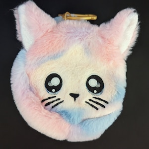 Può includere: Un giocattolo di peluche soffice a forma di gatto rosa e blu con occhi e baffi ricamati. Il giocattolo ha un anello dorato attaccato alla parte superiore della testa.