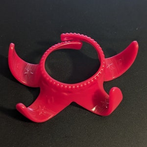 May include: A bright pink plastic egg holder with four prongs. The holder has a beaded rim and a star design on each prong.