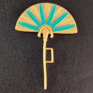 Puede incluir: Un abanico de plástico dorado con un diseño turquesa. El abanico tiene un mango con una parte superior decorativa.
