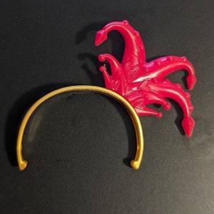 May include: A gold headband with a red plastic jester hat attached. The hat has a pointed tip and multiple curled points.