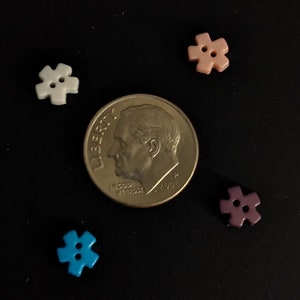 May include: Four plastic buttons in different colors: white, pink, purple, and blue. Each button has a six-pointed star shape with two holes in the center. The buttons are arranged around a US dime coin.