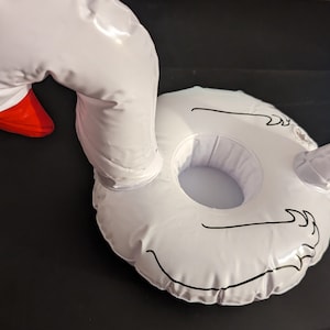 May include: White inflatable swan-shaped drinks holder with a red beak and black detailing. The swan has a circular base with a cup holder in the centre. The swan's neck curves upwards, and the head is tilted to the side.