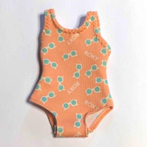 Puede incluir: Un bañador de una pieza naranja con un estampado de gafas de sol azul claro y la palabra "ROXY" impresa. El bañador tiene un ribete festoneado alrededor del cuello y las sisas.