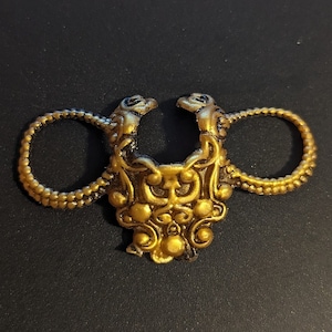 以下が含まれることがあります： 装飾的なデザインの金色の装飾金属リング。リングは、中央の装飾要素を備えた、互いに向き合った2つの様式化されたドラゴンの頭を特徴としています。リングには2つの指ループがあり、装飾品である可能性があります。