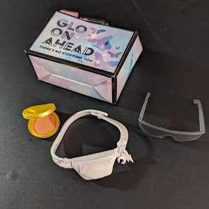 May include: A set of accessories including a small box with the text "GLOW ON AHEAD" and butterfly graphics, a gold compact, a silver fanny pack, and clear plastic glasses. The box also has the text "THERE'S NO STOPPING YOU".
