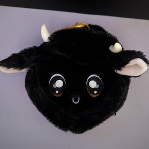 Può includere: Giocattolo di peluche nero con una texture soffice, a forma di agnello con corna bianche e orecchie rosa. Il giocattolo ha occhi ricamati e una bocca sorridente.