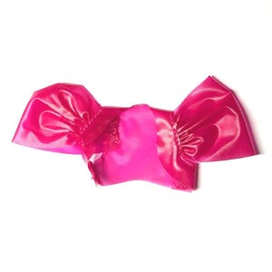 May include: A bright pink satin bow, likely for hair or clothing embellishment, lies flat against a white background. The bow has a central section and two flared, ruffled wings. The fabric has a slight sheen.