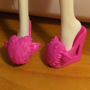 Könnte beinhalten: Ein Paar hellrosa Puppenschuhe. Die Schuhe haben einen Keilabsatz und ein strukturiertes, stacheliges Design auf der Vorderseite. Die Schuhe sind für den Fuß einer Puppe konzipiert.