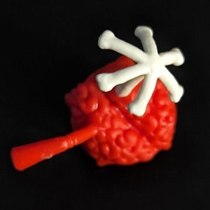 May include: A red and white plastic toy with a skull-shaped head and a bone-shaped handle. The toy is designed to look like a brain with a bone sticking out of it.