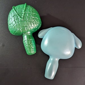 May include: Two plastic toys, one green and one blue. The green toy is shaped like a small, round, handheld drum with a beaded strap. The blue toy is shaped like a small, round, handheld drum with a smooth surface.
