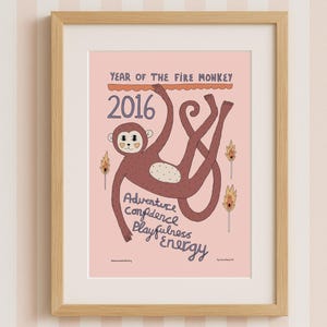 Może przedstawiać: Oprawiony wydruk z ilustracją małpy z kreskówki. Wydruk jest w drewnianej ramie i ma różowe tło z napisem "Rok Ognistej Małpy" i "2016". Małpa jest brązowa z białym brzuchem i słowami "Przygoda, Pewność siebie, Zabawa, Energia".