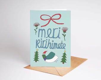 Op de afbeelding: Lichtblauwe wenskaart met een rode strik, tekst en illustraties. De kaart toont de woorden "meri Ririhinete" in blauw, met een duif met een kerstmuts, twee kerstbomen en bloemen. De kaart ligt op een bruine envelop.