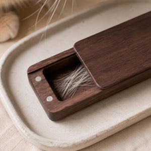 Puede incluir: Una caja deslizante rectangular de madera marrón oscuro con una tapa parcialmente abierta. El interior de la caja contiene un pequeño grupo de fibras finas y claras. Se observan dos pequeños imanes metálicos circulares de color plateado.