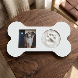 May include: White bone-shaped pet memorial frame. The frame holds a photo of a dog and a paw print impression. The frame is on a dark wood surface.