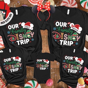 Puede incluir: Camisetas negras a juego para la familia con el texto "OUR Family DISNEY TRIP" en rojo, verde y blanco. Las camisetas tienen nombres personalizados debajo de un gorro de Pap&aacute; Noel. Tambi&eacute;n se ven una diadema de Minnie Mouse y una bolsa de mano.