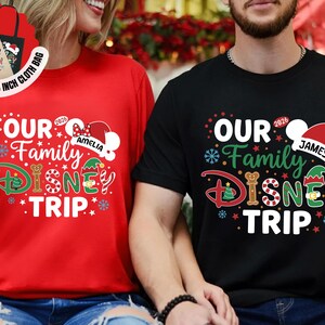 Puede incluir: Camisetas rojas y negras con el texto "Our Family Disney Trip" en un dise&ntilde;o festivo. La camiseta roja tiene el nombre "Amelia" y la camiseta negra tiene el nombre "James". La imagen tambi&eacute;n muestra una bolsa de tela gratuita de 38x38 cm.
