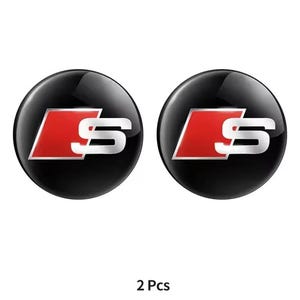 Puede incluir: Dos emblemas de coche negros y circulares con un logotipo "S" plateado y rojo. Los emblemas son brillantes y tienen una superficie reflectante. El texto "2 Pcs" está en la parte inferior de la imagen.