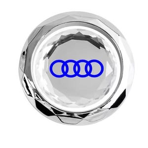 May include: A clear, faceted crystal car emblem with a silver-colored rim. The emblem features the blue Audi logo, consisting of four interlocking rings. The design is circular and has a reflective surface.