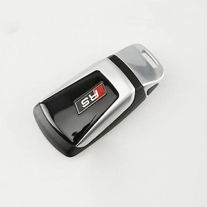May include: A car key fob with a black and silver design. The key has a rectangular shape with a silver top and a black center section. The center features a red and white "RS" logo.