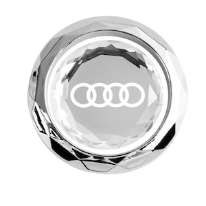 May include: A chrome-colored, faceted button with the Audi logo in white. The button has a circular shape with a clear, reflective center. The design is modern and sleek, suitable for automotive accessories.