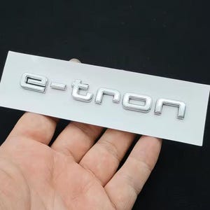Puede incluir: Emblema plateado "e-tron" sobre un respaldo rectangular blanco. El emblema tiene un acabado metálico y lo sostiene una mano sobre un fondo oscuro. Las letras están en una fuente moderna, sans-serif.