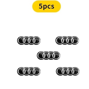 Puede incluir: Cinco insignias de emblema de coche negras y plateadas con el logotipo de Audi, que presentan cuatro anillos entrelazados. El texto "5pcs" está en un rectángulo amarillo en la parte superior de la imagen.