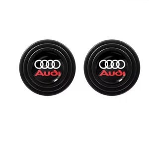 May include: Two black, circular car door protectors. Each features the Audi logo in white and the Audi name in red. The protectors have a ridged design and are set against a white background.