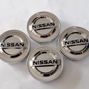 Peut inclure: Quatre enjoliveurs chromés avec le nom de marque "NISSAN" en lettres noires. Les enjoliveurs sont circulaires avec un centre surélevé et une surface brillante et réfléchissante. Le nom de la marque est centré sur une barre rectangulaire.
