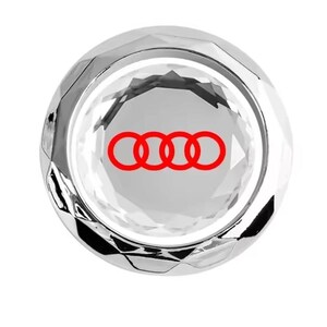 Op de afbeelding: Een zilverkleurig auto-accessoire met een rood Audi-logo in het midden. Het logo bestaat uit vier in elkaar grijpende ringen. Het accessoire heeft een reflecterend, kristalachtig uiterlijk.
