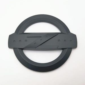 May include: A matte black emblem featuring a circular outer ring with a rectangular center section. The center has a diagonal design and several small rectangular cutouts. The emblem is likely for automotive use.
