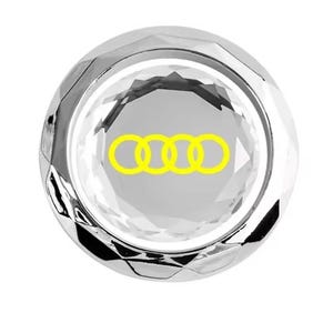 May include: A clear, faceted crystal car emblem with a yellow Audi logo. The emblem is circular and has a shiny, reflective surface. The Audi logo consists of four interlocking rings.
