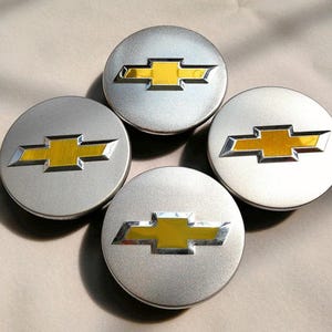 May include: Four silver-colored hubcaps with a yellow and silver Chevrolet logo in the center. The hubcaps are round and have a metallic finish. The Chevrolet logo is a stylized bow tie.