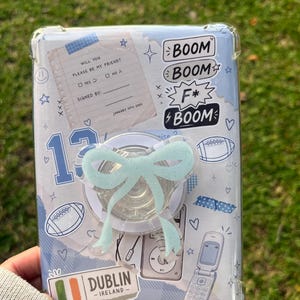Op de afbeelding: Een doorzichtig telefoonhoesje met een collage van lichtblauwe en witte afbeeldingen, waaronder een vriendschapsbriefje, voetballen, een klaptelefoon en een sticker van Dublin, Ierland. Een lichtblauwe glitterende strikvormige telefoonhouder is in het midden bevestigd.
