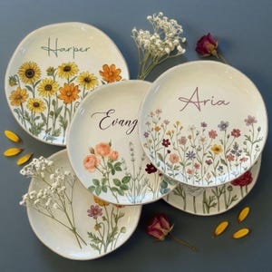 Op de afbeelding: Een verzameling witte keramische borden met bloemmotieven en gepersonaliseerde namen. Eén bord toont zonnebloemen, een ander rozen en een derde wilde bloemen. De namen "Harper", "Evang" en "Aria" staan op de borden.