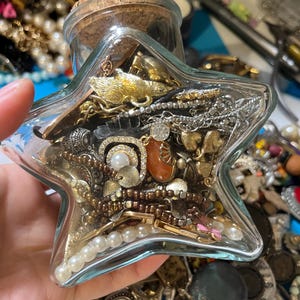 May include: A clear, star-shaped glass jar filled with an assortment of jewelry, including necklaces, earrings, and brooches. The jar is sealed with a cork stopper. The jewelry pieces are in various colors, including gold, silver, and pearl.